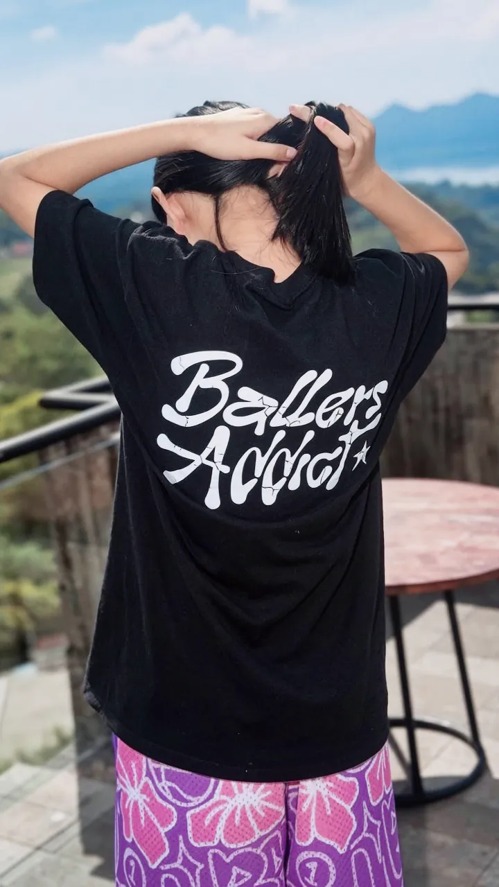 A black tee with a white Ballers Addict back print over Purple Urban Doodle Shortpants shorts, seen from behind on a terrace above a valley.