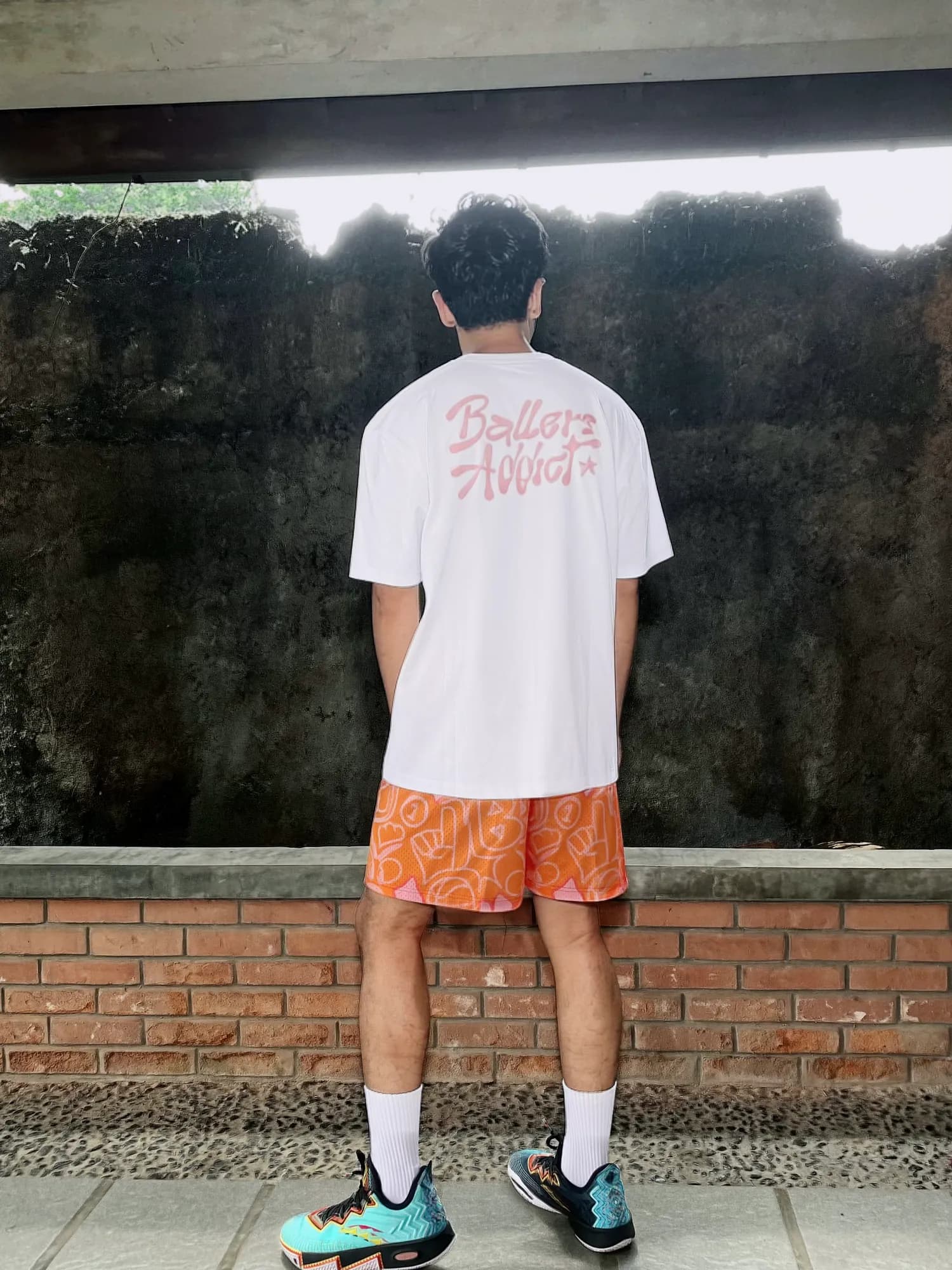 Orange Urban Doodle Shortpants shorts and a white tee with a pink Ballers Addict back print, seen from behind against a mossy wall.