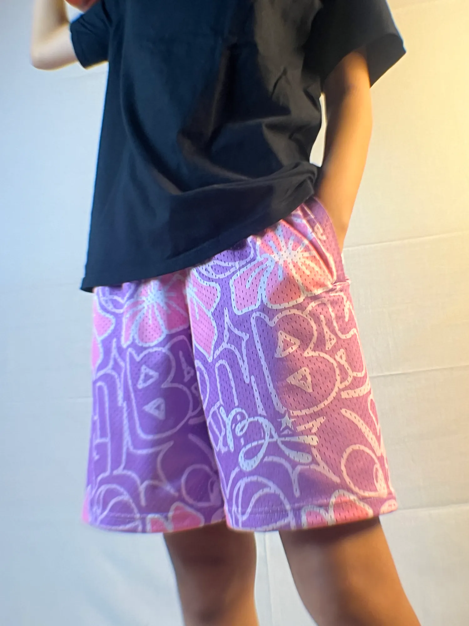 Purple Urban Doodle Shortpants shorts, lilac mesh printed with pink hibiscus, worn with a black tee and a hand in the pocket.